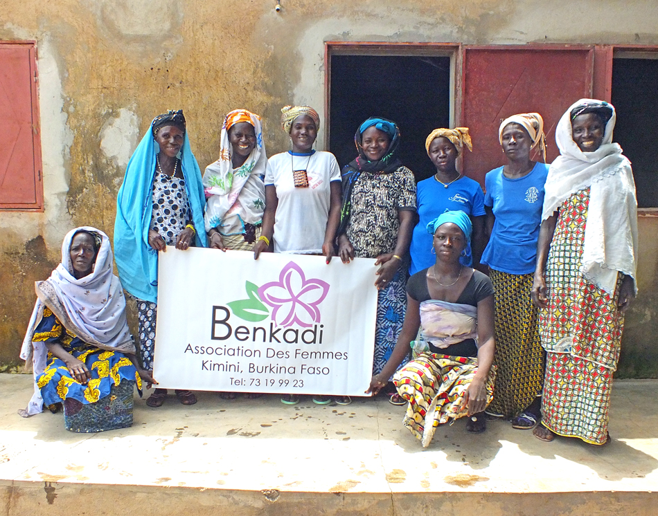 A group of 9 women stand together proudly, holding up a banner for their new business called Benkadi in Burkina Faso
